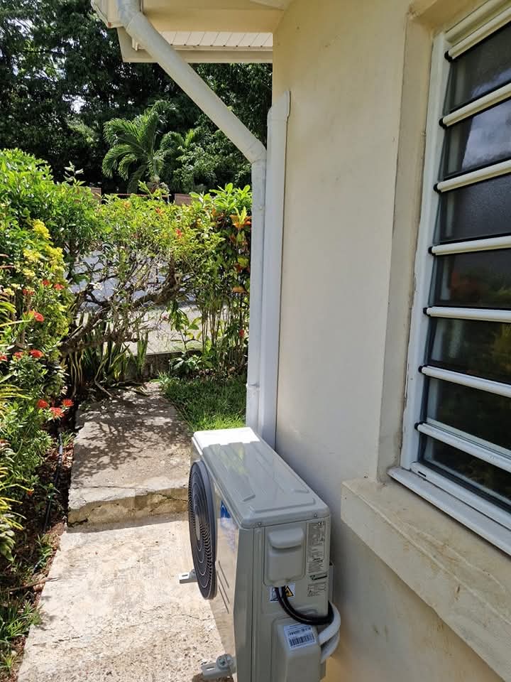Installation Midea 9 000 BTU à Ducos : unité extérieure posée au sol sur une allée en béton, adossée à la façade et abritée par l'avant-toit – Ducos, Martinique – LD CLIM 972