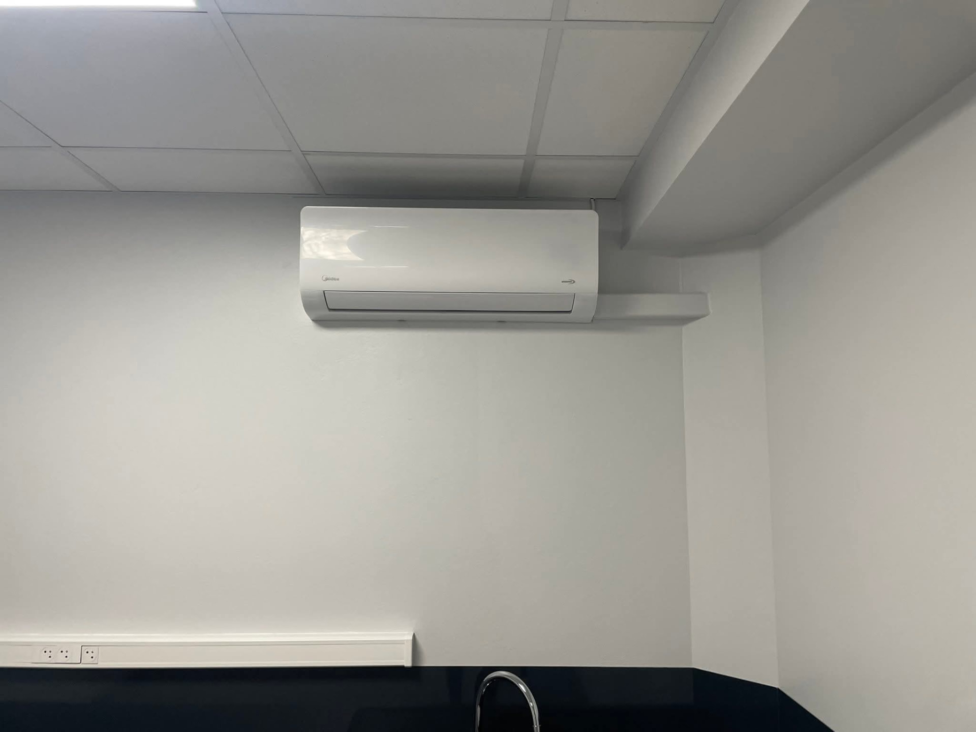 Climatiseur mural Midea installé dans le coin cuisine d'une entreprise au Lamentin, liaisons protégées par une goulotte blanche au-dessus du point d'eau – Lamentin, Martinique – LD CLIM 972