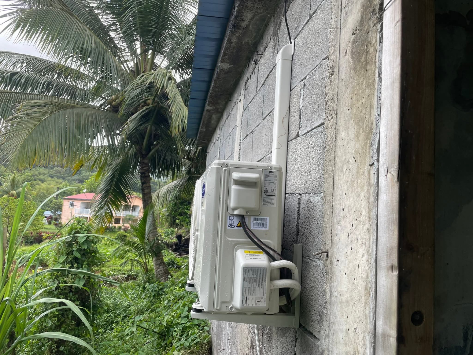 Remplacement 9k BTU – Le Gros-Morne – climatisation Martinique Remplacement 9 000 BTU au Gros-Morne : unité extérieure posée sur support mural contre un mur en parpaing, au milieu de la végétation tropicale – Le Gros-Morne, Martinique – LD CLIM 972